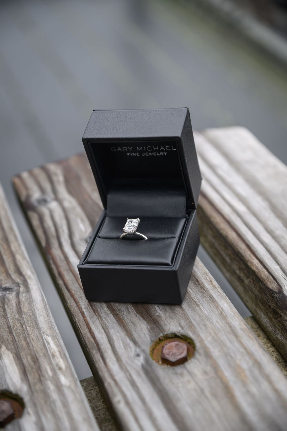 Black ring box containing a diamond engagement ring sits on weathered wooden planks.