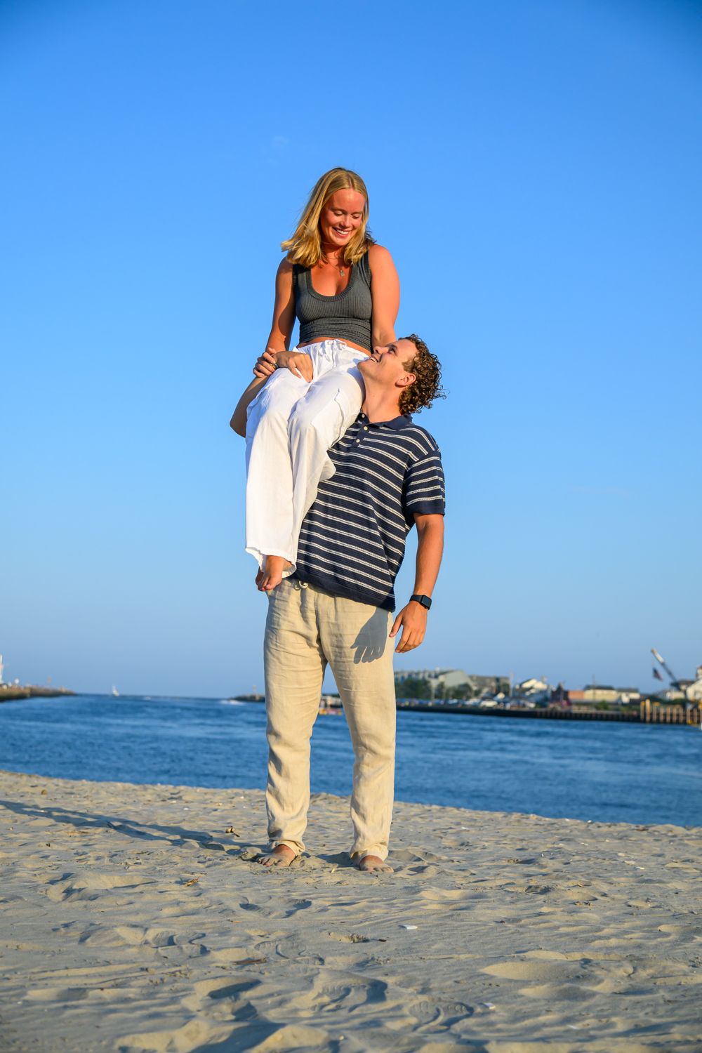 A couple plays and laughs together on a sandy beach with blue ocean and clear sky in the background.