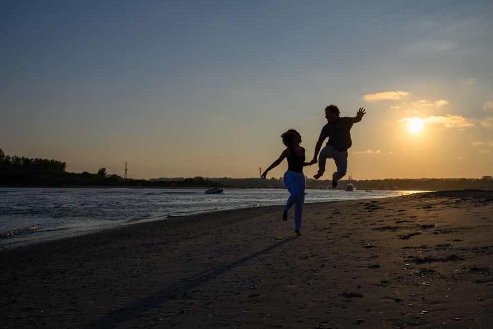 Silhouettes of people jumping joyfully on a beach at sunset with golden sun peeking through clouds.