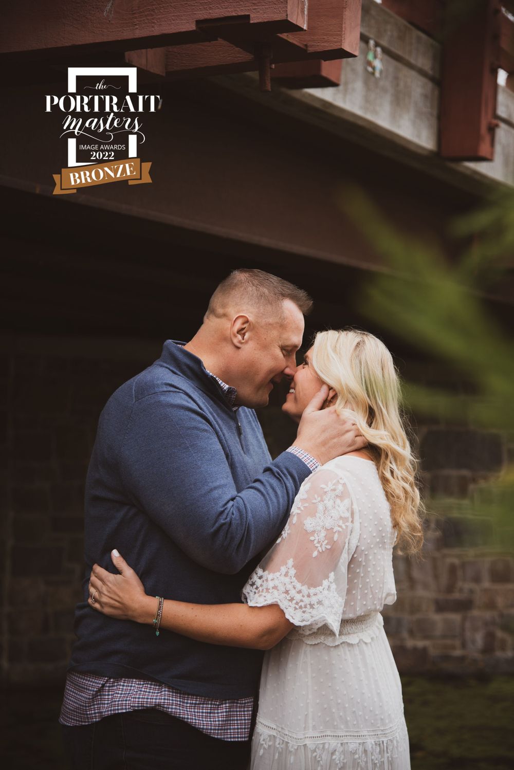 Intimate couple's portrait under rustic bridge showing tender embrace.