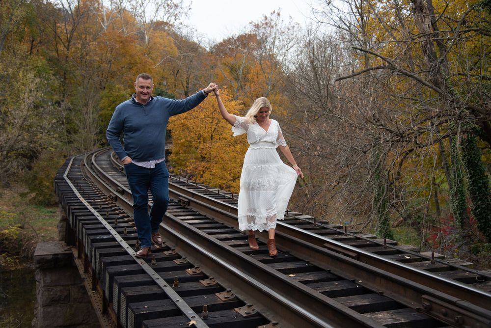 A couple playfully poses for photos on rustic train tracks surrounded by autumn foliage.
