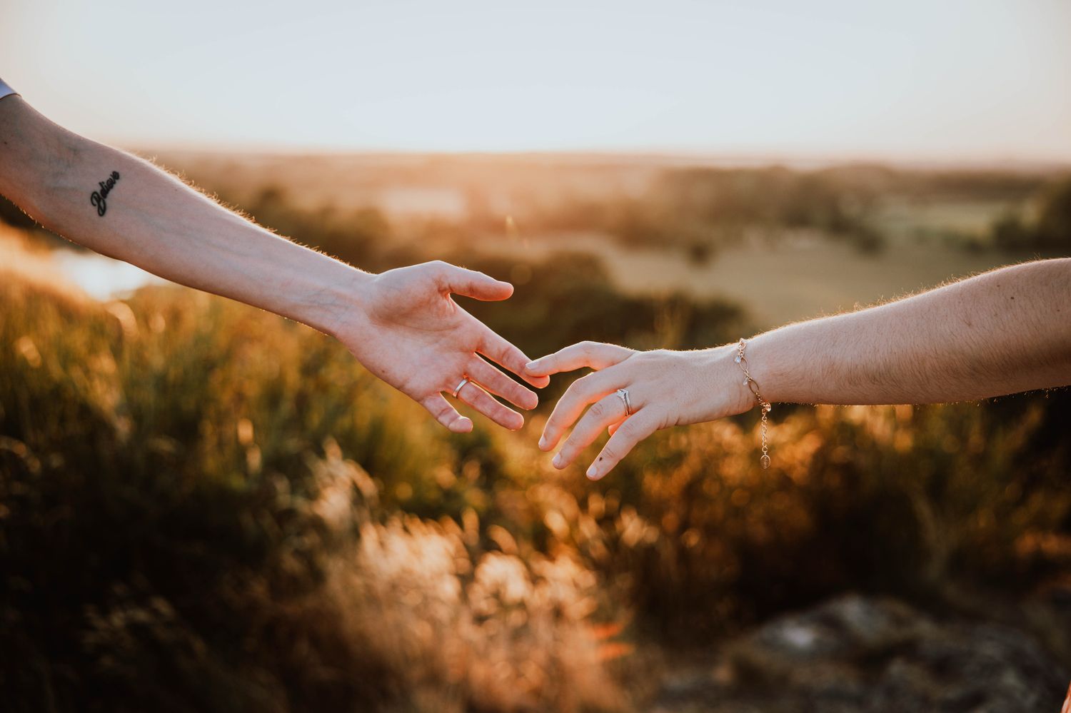 Two hands reach towards each other against a warm sunset backdrop with golden light filtering through tall grass.