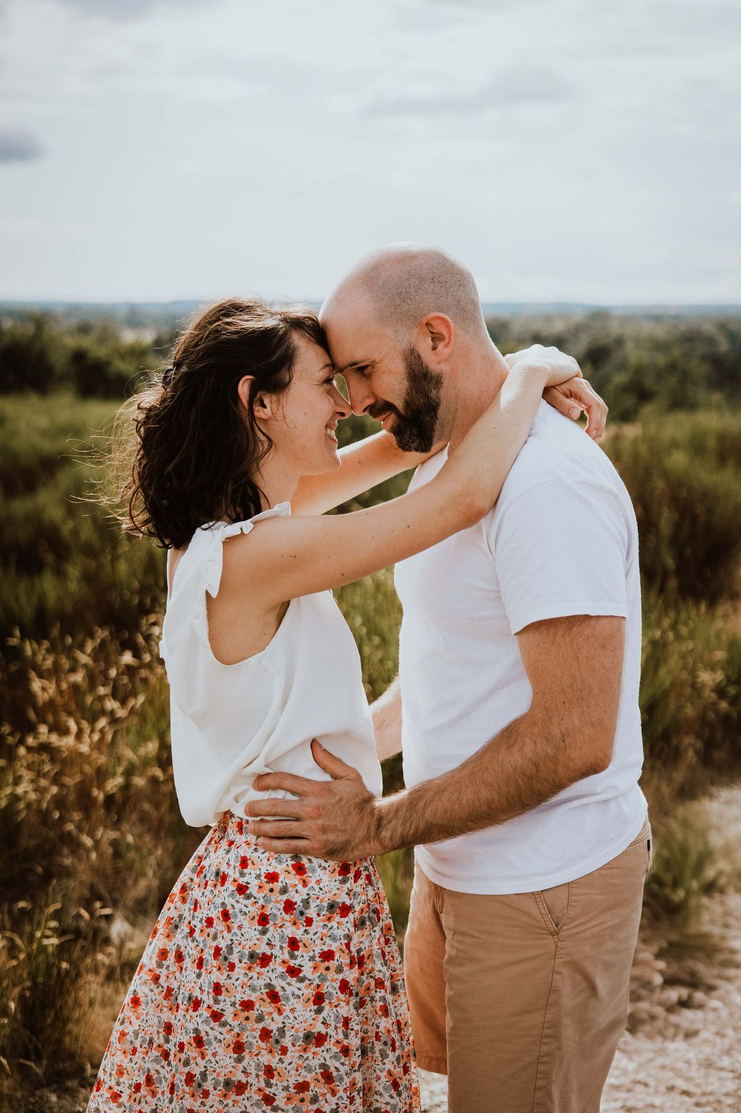 A romantic couple embraces in a sunny outdoor field while wearing white tops and neutral tones.