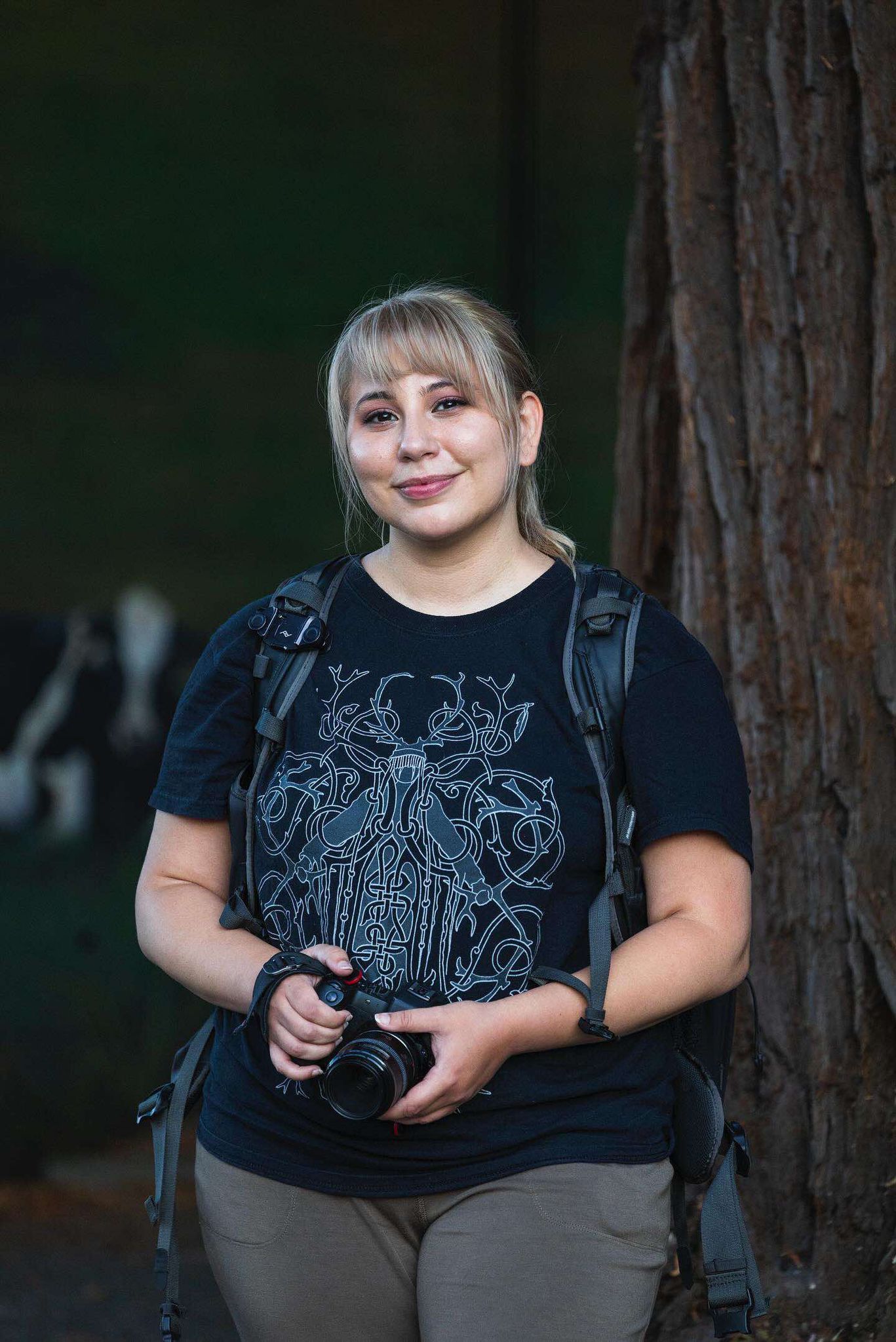 Photographer with camera wearing graphic t-shirt stands by tree trunk.