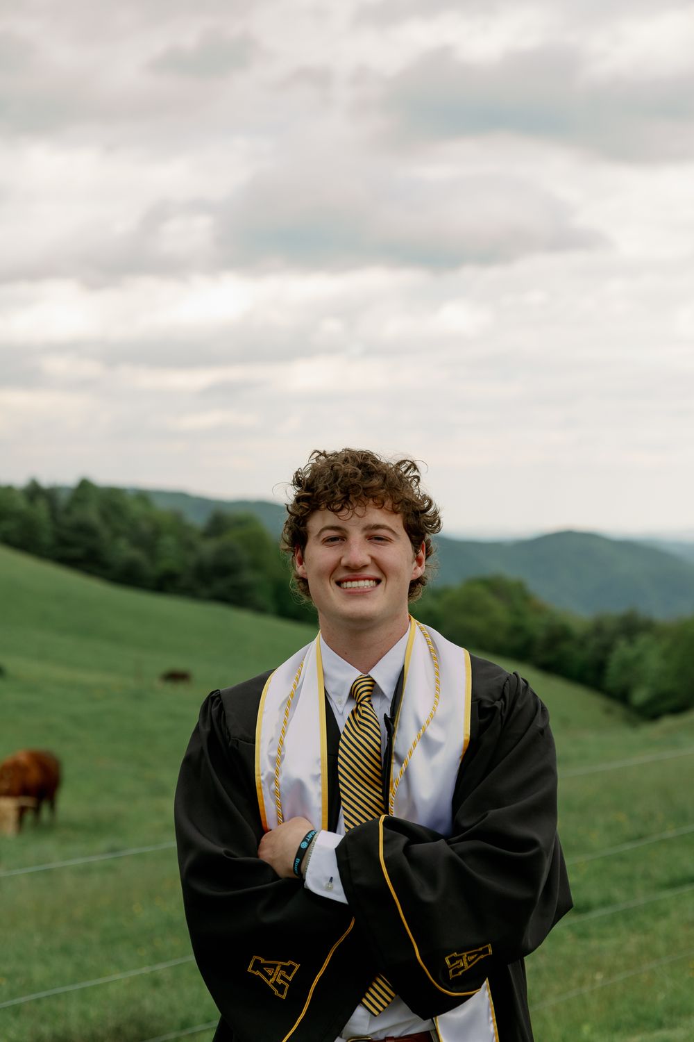 Graduate in black cap and gown poses in front of mountain landscape with cows grazing in background.