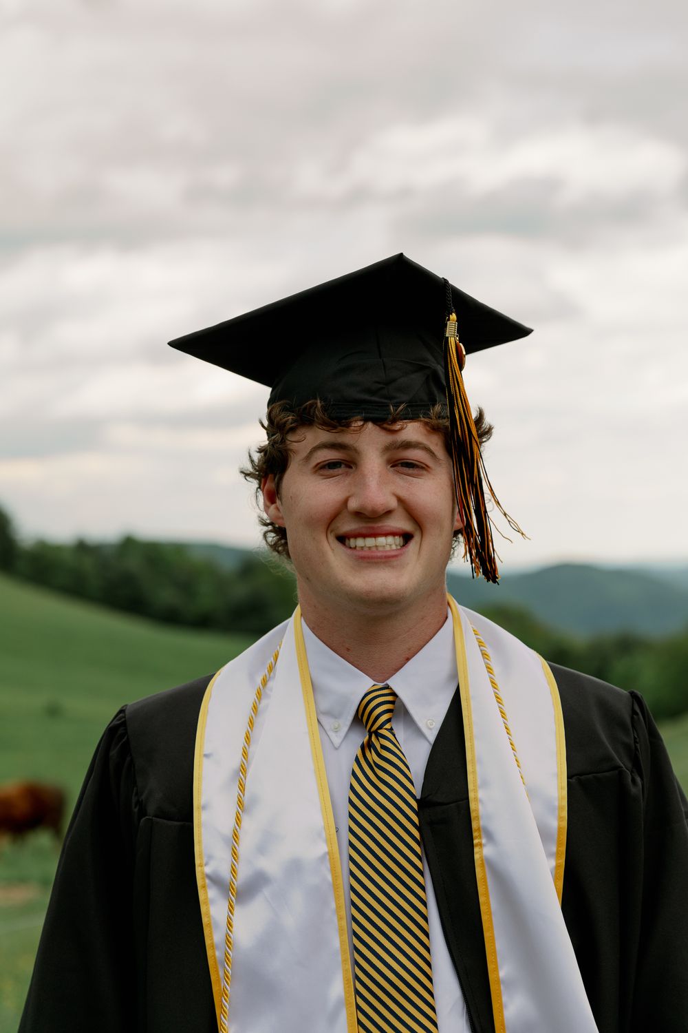 Graduate wearing black gown and striped tie smiles while posing in cap and tassel.