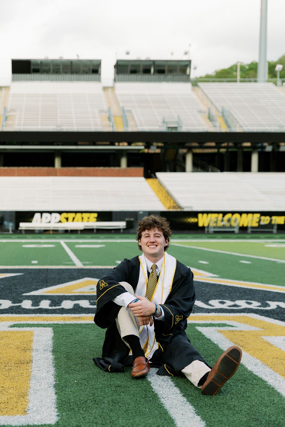 Graduate in academic regalia lying on football field with stadium seating in background.