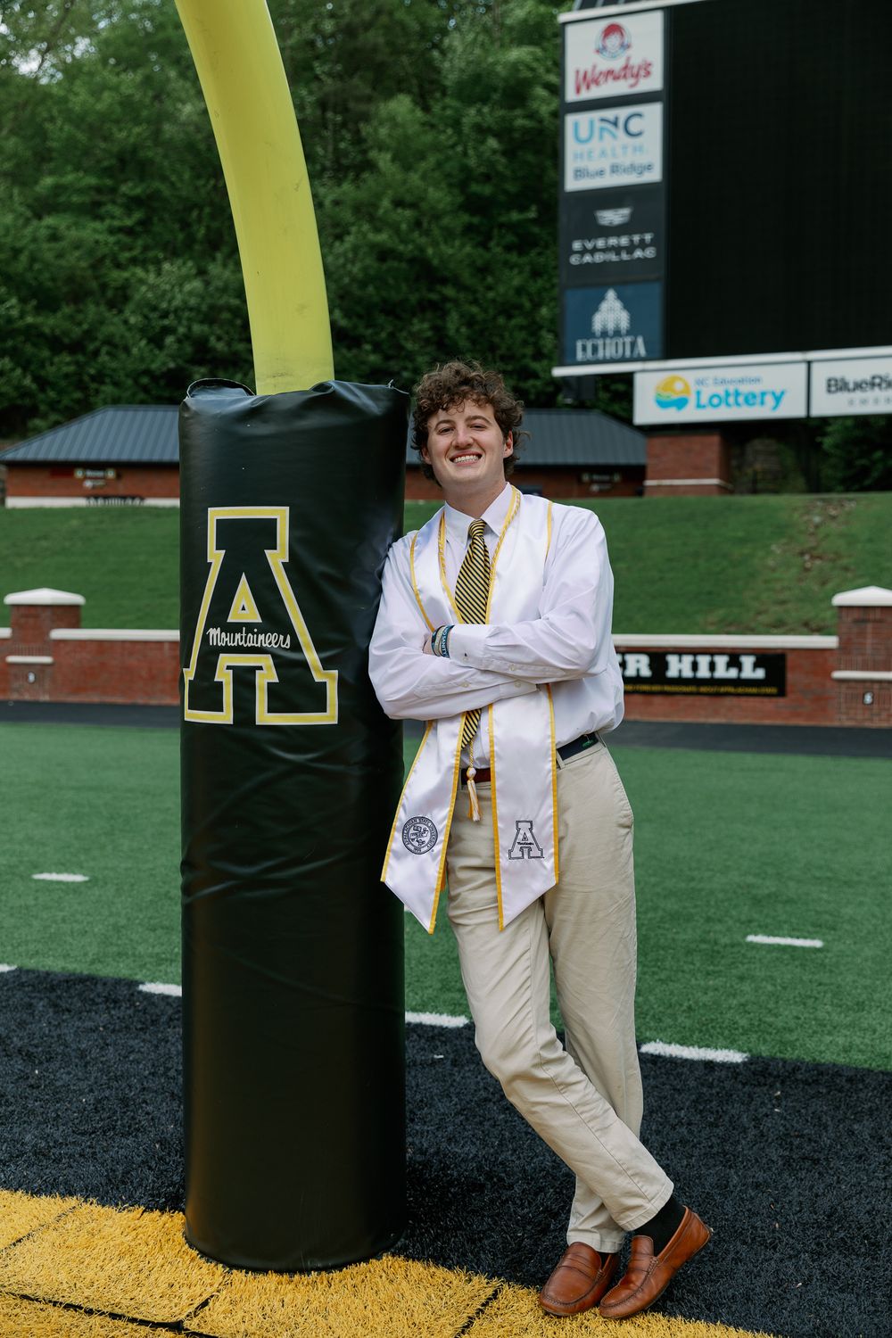 Graduate poses near football field goal post wearing dress shirt and striped tie.
