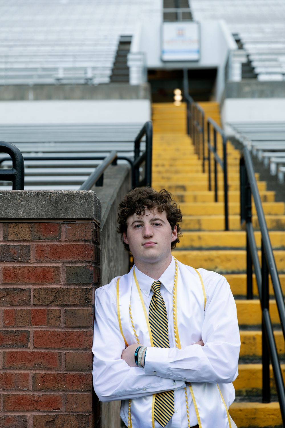 Graduate in white shirt and striped tie poses against brick wall with stadium bleachers.