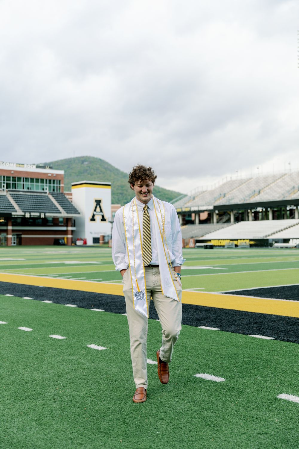 Graduate walks across football field in formal attire with mountains visible in background.