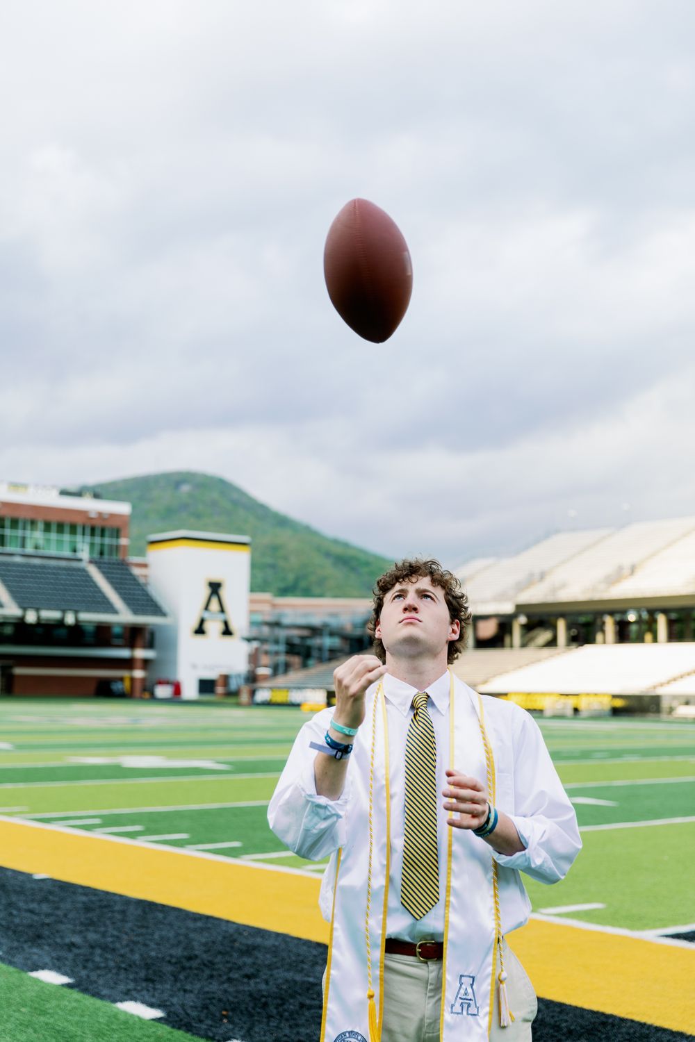 Graduate in formal attire tosses football in the air on athletic field.