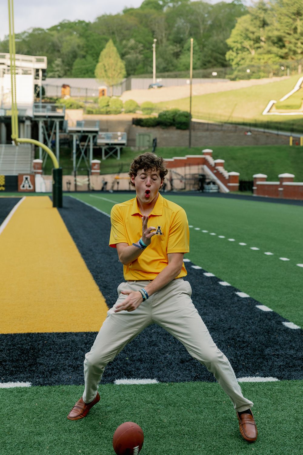 Person in yellow polo shirt plays with football on athletic field.
