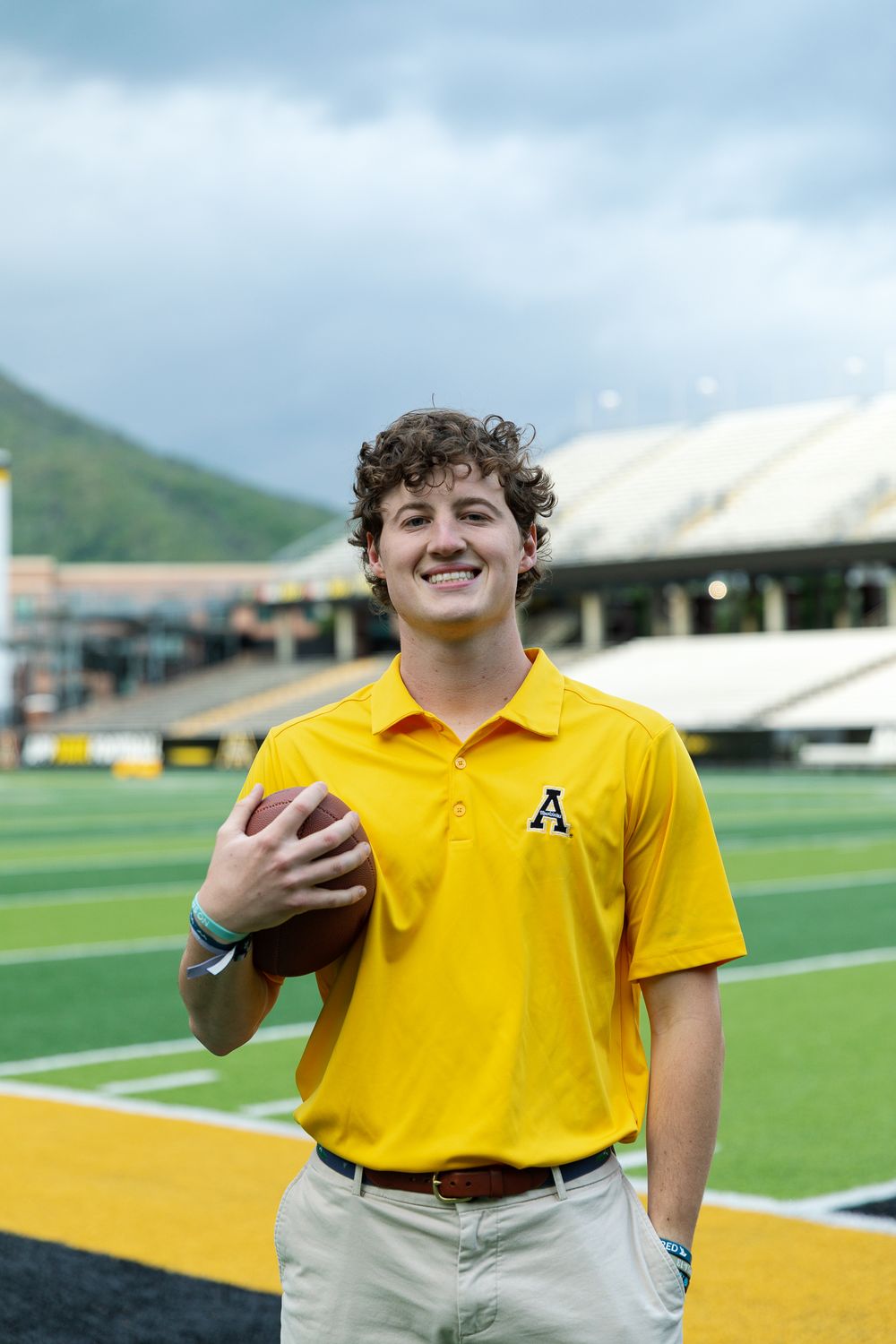 Student in yellow polo shirt poses with football on athletic field with stadium in background.