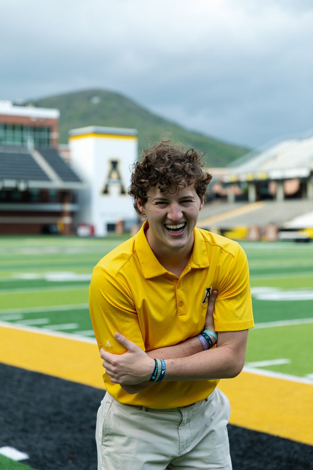 Student in yellow polo laughs while playing with football on athletic field.
