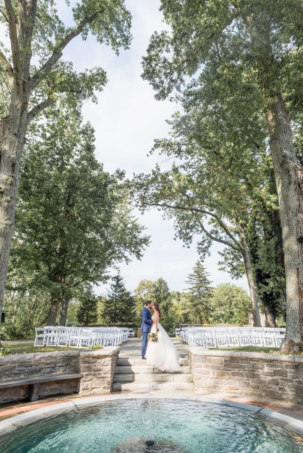 A scenic outdoor wedding ceremony setup with white chairs near a stone fountain surrounded by tall trees.