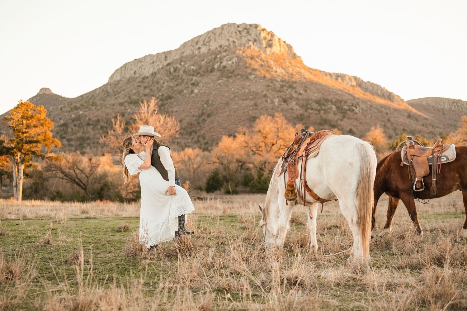 A couple dressed in western attire sharing a kiss next to horses.