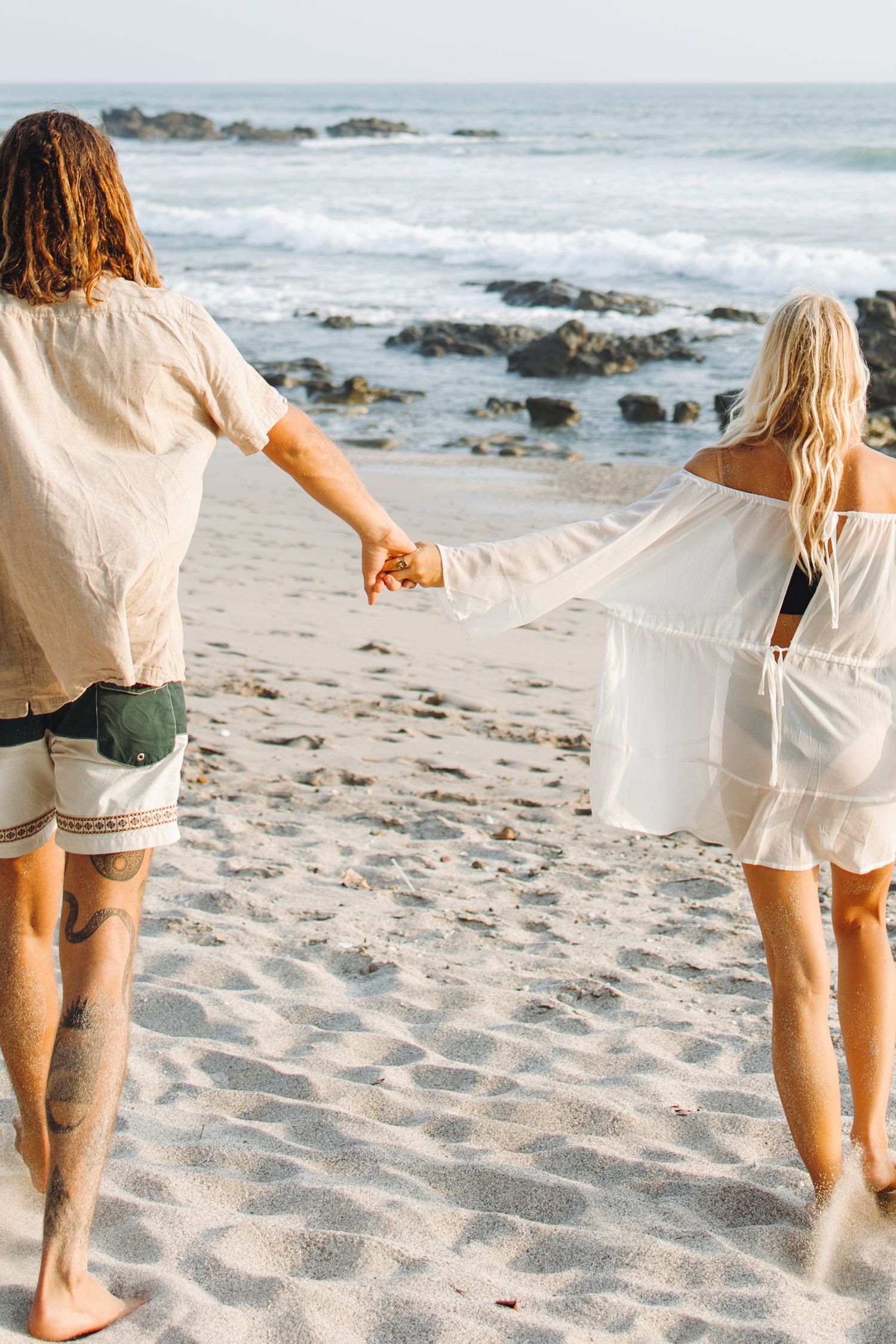 Two people holding hands while walking barefoot on a sandy beach by the ocean.