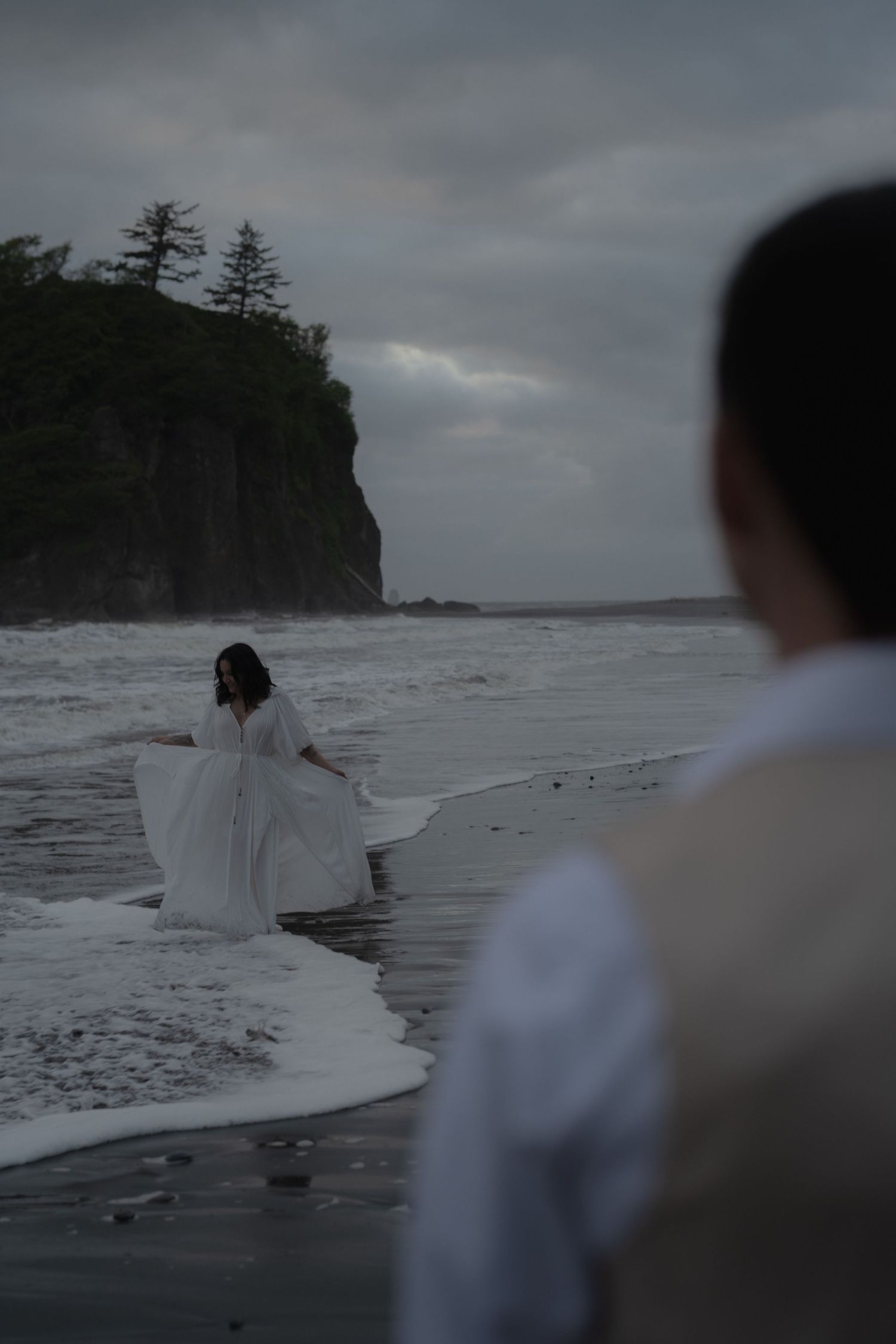 Person in white dress dances on misty beach shore near dramatic cliffs.