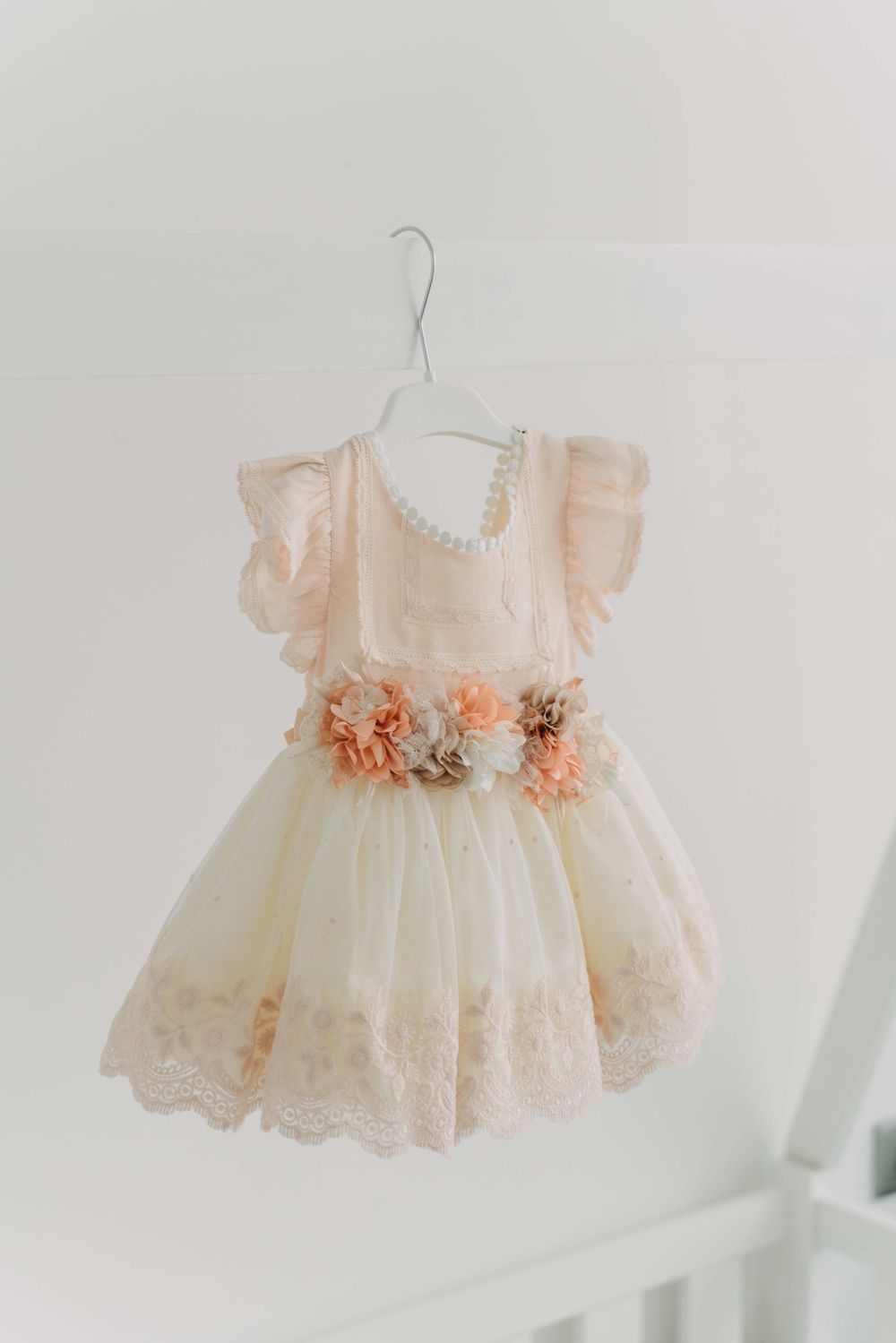 Delicate peach and ivory baby dress with floral details hanging on white hanger.