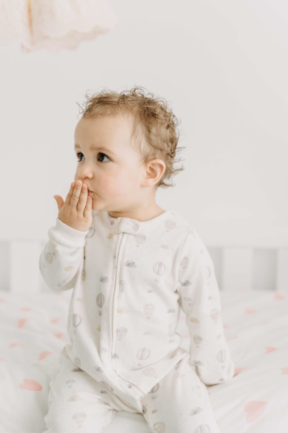 Baby in white sleepwear sitting with hand near mouth.