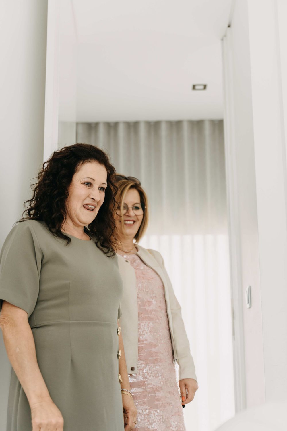 Two women in formal attire stand together smiling in a light-filled room.
