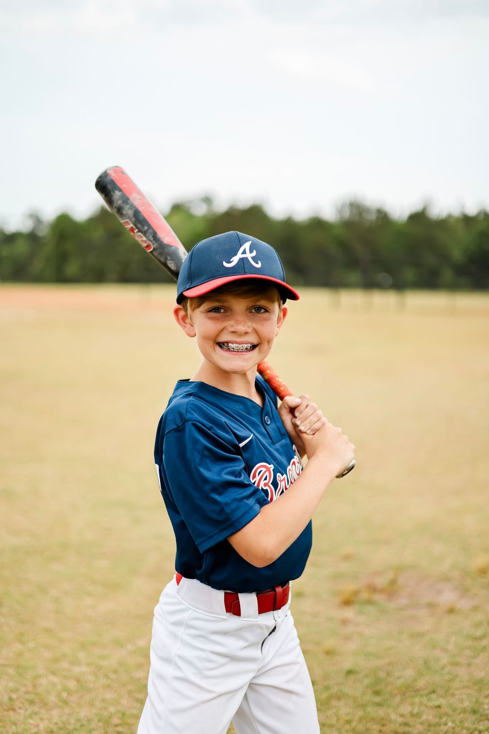Young baseball player in Atlanta Braves uniform poses with bat on sunny field.