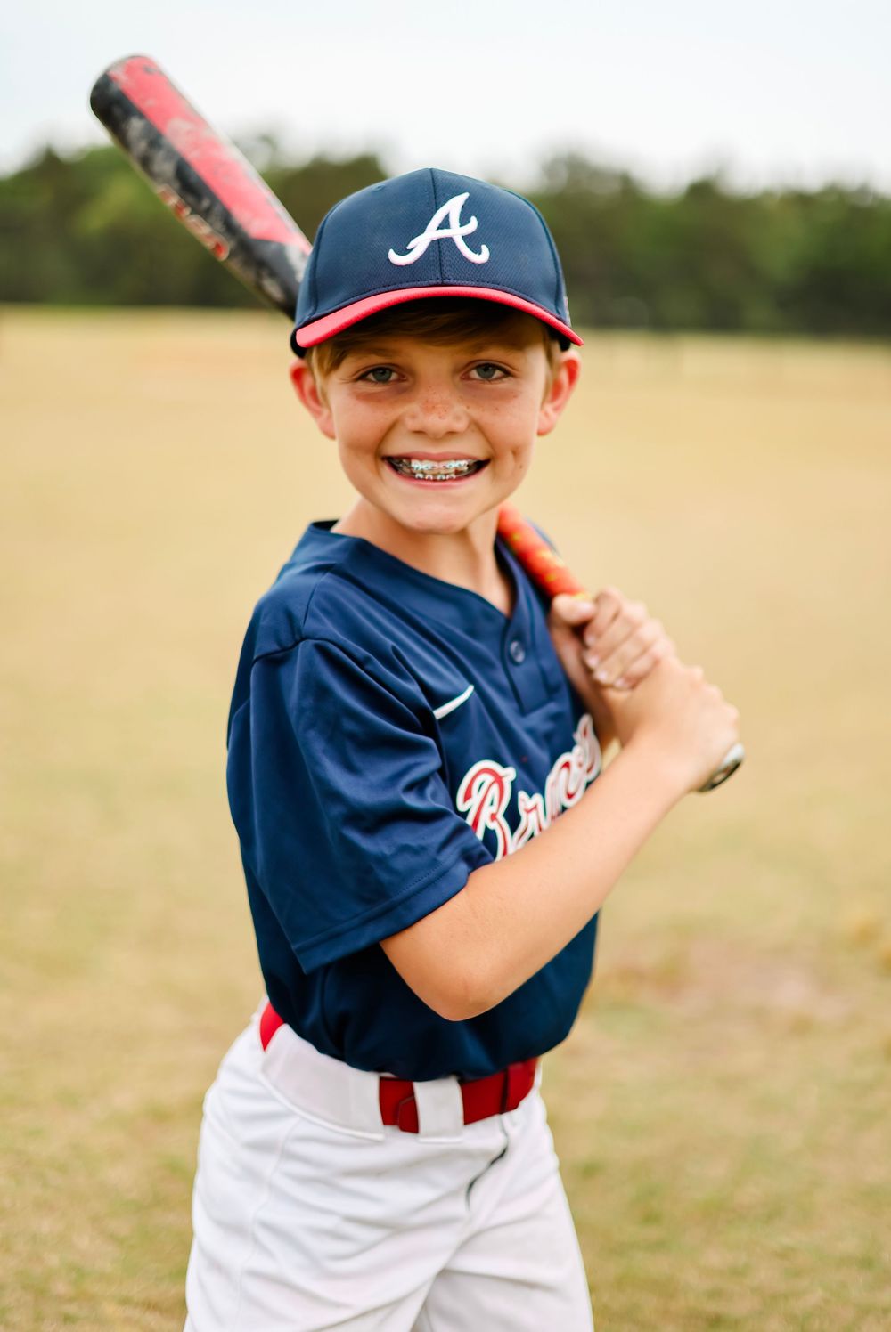 Young baseball player in Atlanta Braves uniform poses with bat on sunny field.