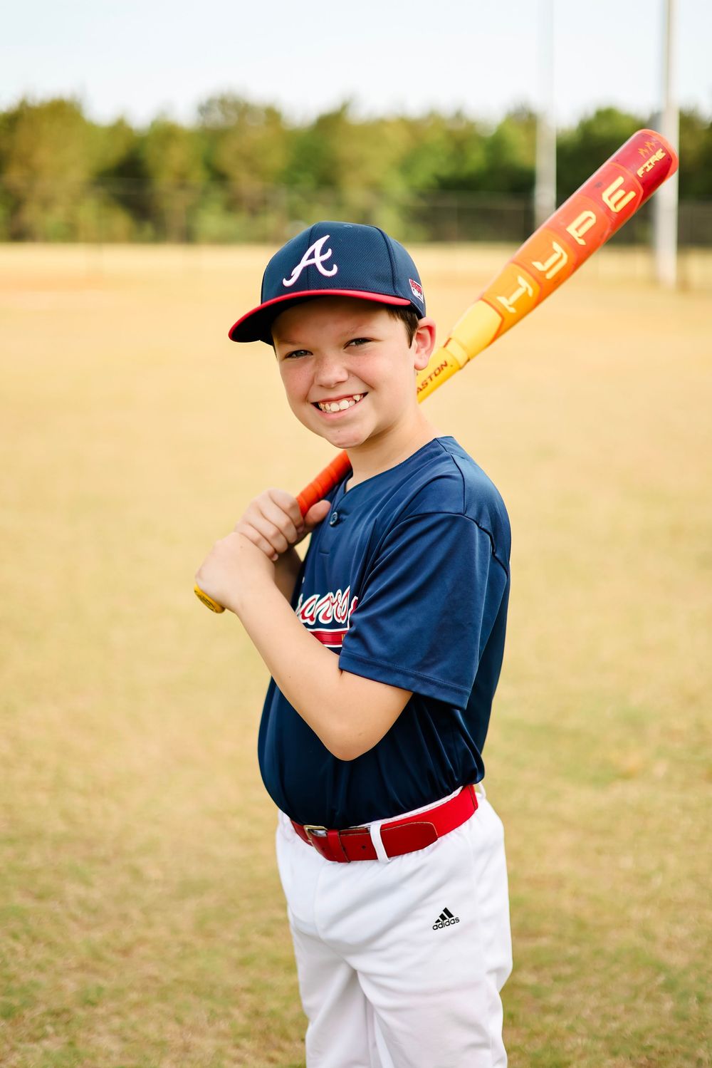 Young baseball player in Atlanta Braves uniform poses with orange bat in grassy field.