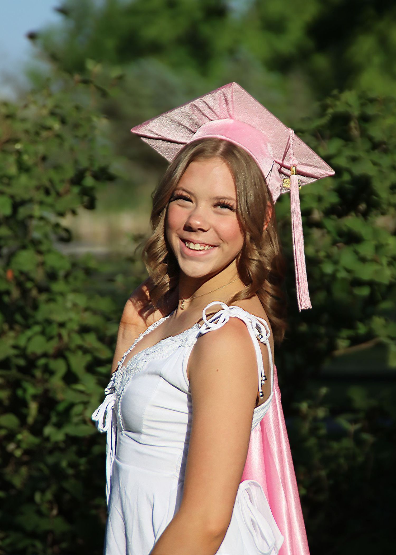 A graduation portrait featuring a pink cap and white dress against a green leafy background.