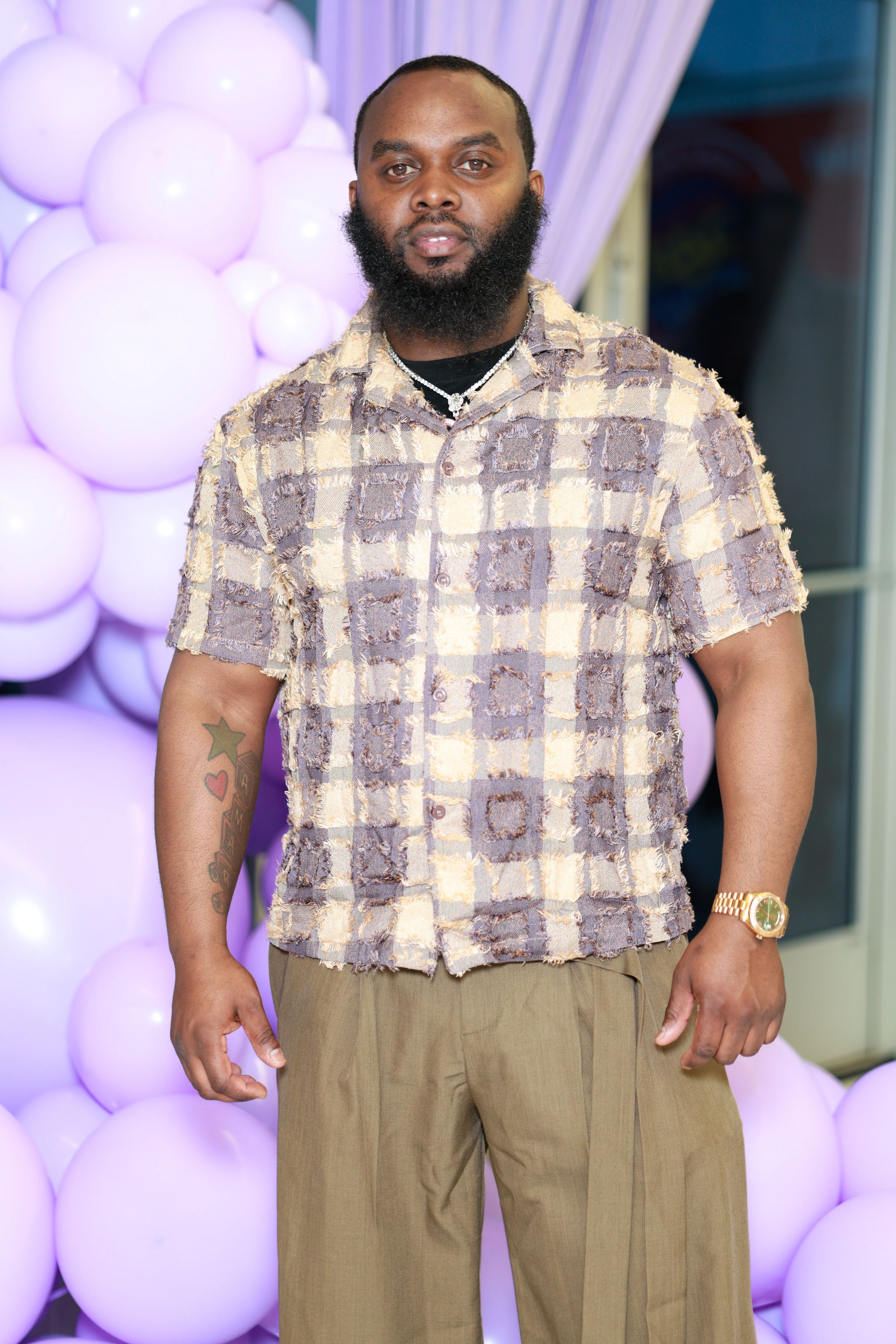 Man in plaid textured shirt poses in front of purple balloon backdrop at celebration.