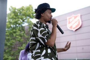 A performer in a patterned shirt and cap holds a microphone while speaking at an outdoor event with a Salvation Army logo visible.
