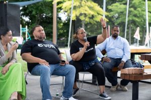 A panel discussion takes place outdoors with several speakers seated in front of American flag decorations.
