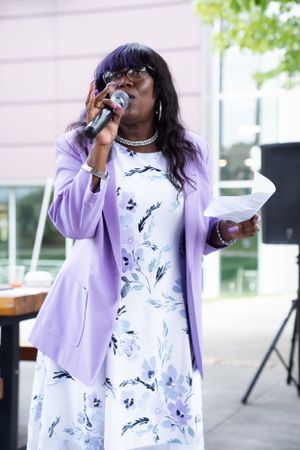 A performer in a lavender blazer and floral dress sings into a microphone at an outdoor event.