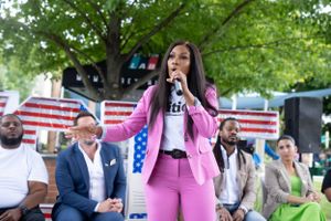 A speaker in a bright pink outfit addresses an audience at an outdoor political event with American flags.