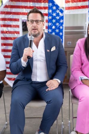 A speaker in a navy suit addresses an audience at an outdoor event with American flags in the background.