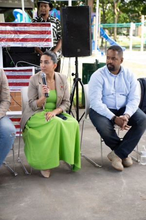 A speaker in green dress addresses audience while others listen during outdoor panel discussion.
