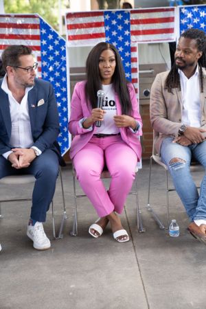Three panelists engage in discussion at outdoor event with American flags displayed behind them.