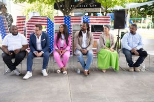 People sit on chairs against an American flag backdrop at an outdoor event.