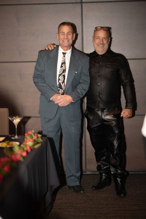Two men posing together at formal event with cocktail table and flowers in background.