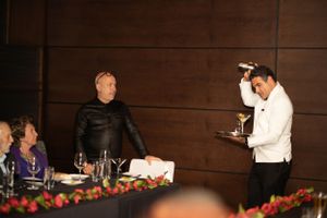 Server in white jacket presenting drinks at formal dinner with decorative floral centerpieces.