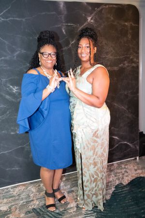 Two guests pose together against a marble wall at an elegant event.
