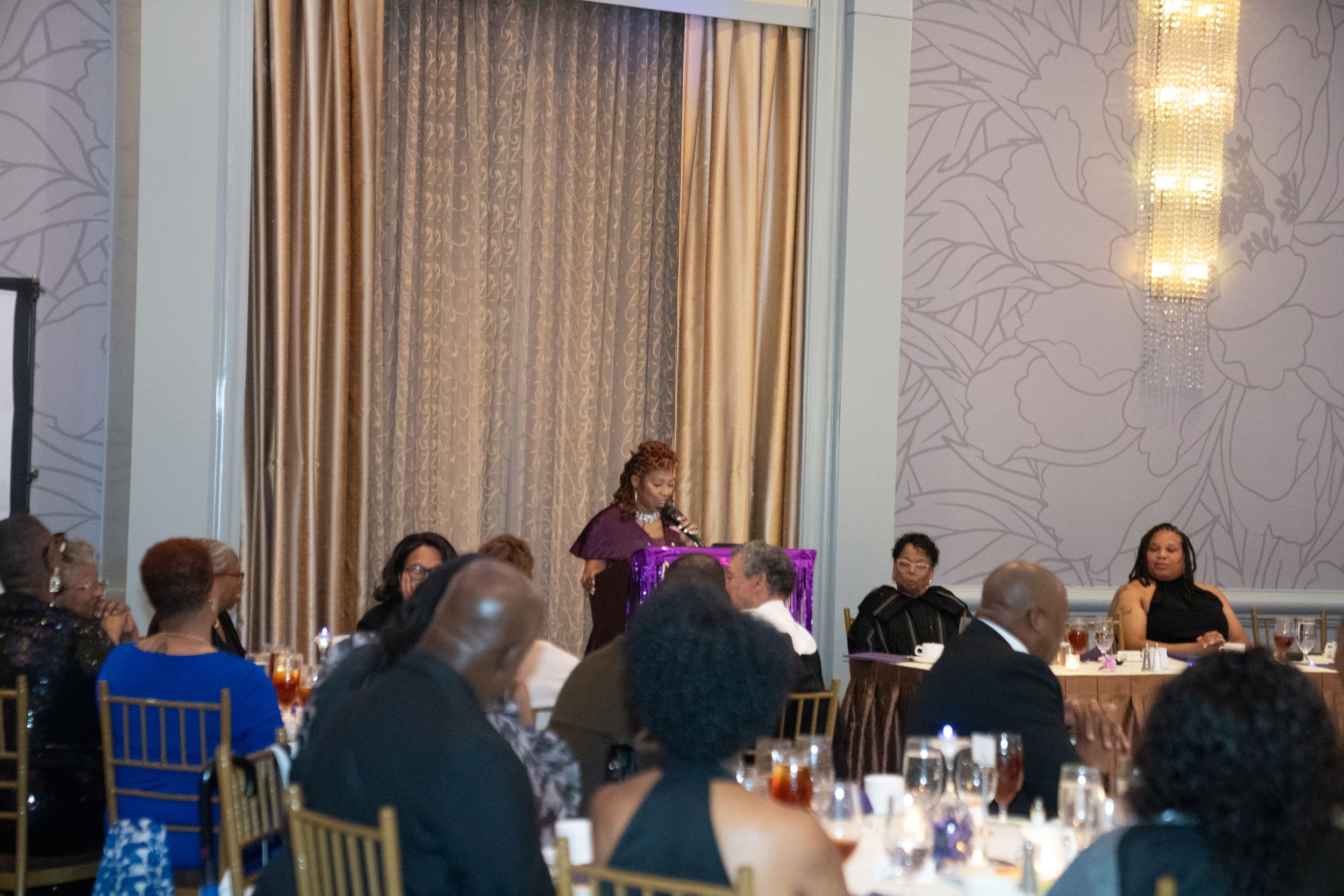 Speaker addresses guests during formal dinner event in elegant ballroom setting.