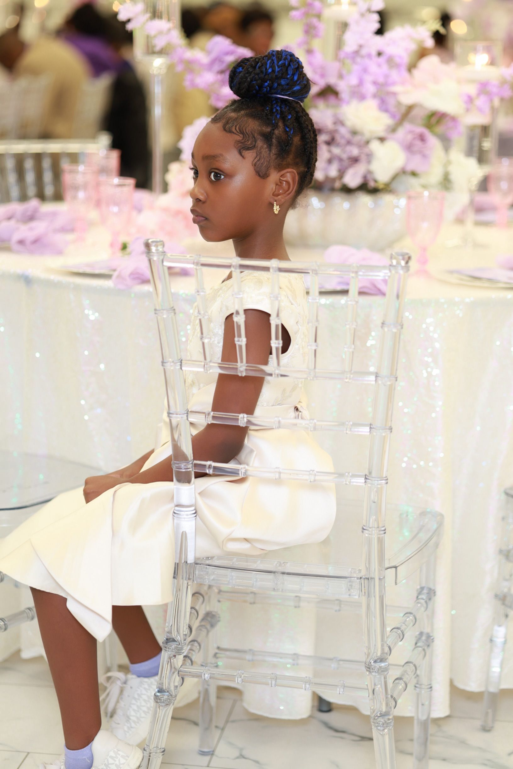 Young girl in white dress sits on clear chiavari chair at elegant lavender-themed event.