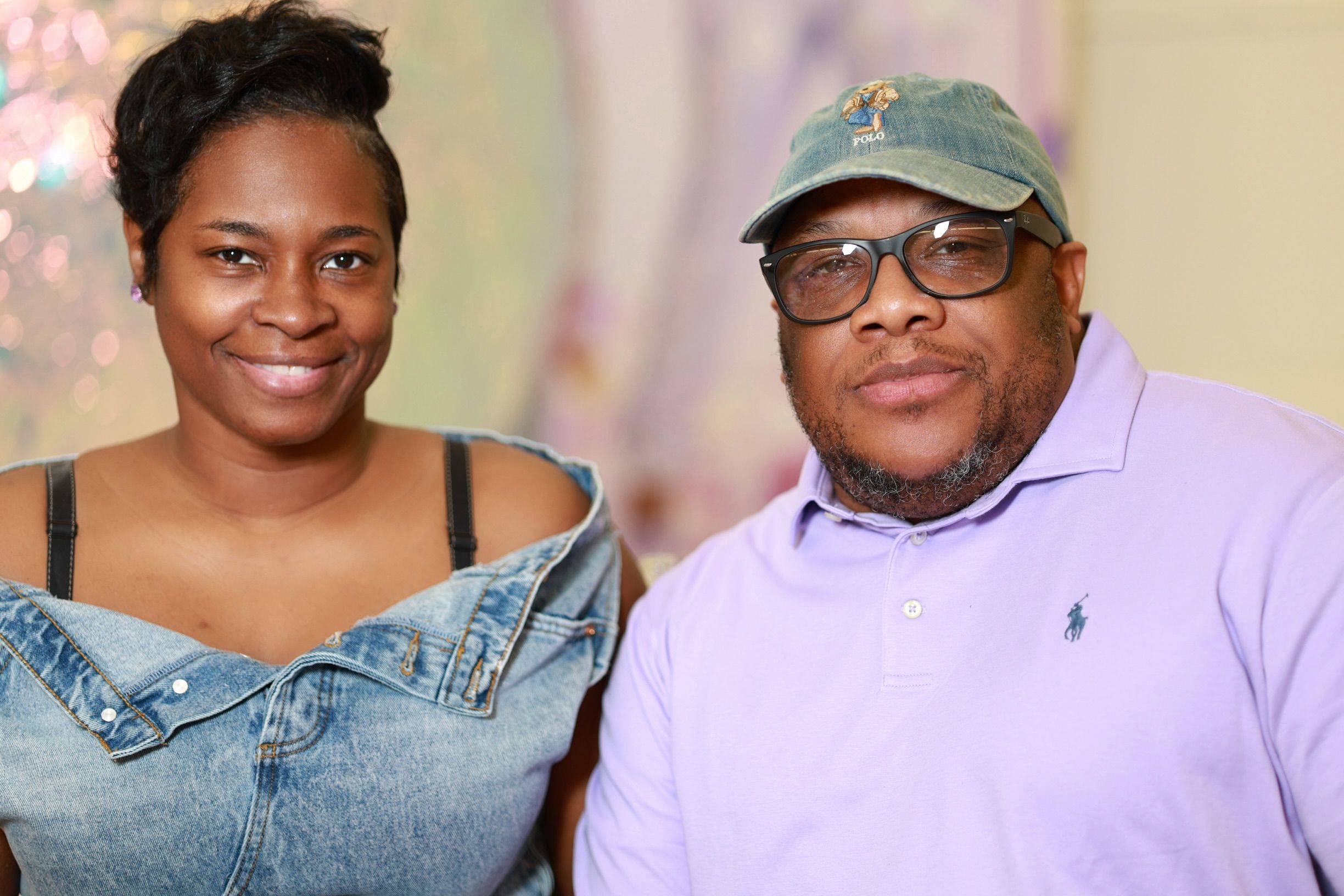 Two people smiling and posing together, a woman in a denim top and a man in a purple polo shirt and green cap.