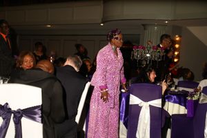A person in a sparkly pink evening gown walks through an elegant reception hall with purple and white decorated chairs.