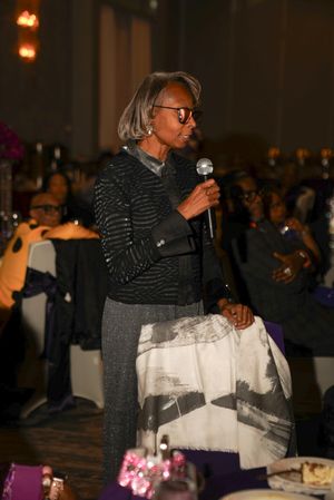 Speaker with microphone at formal event wearing striped sweater and glasses.