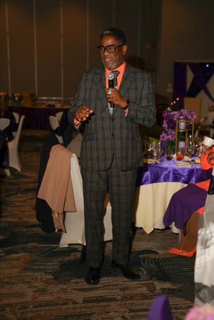 A person in a plaid suit walks across a carpeted ballroom with purple and white decorated tables.