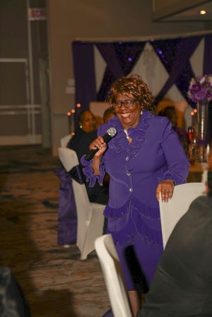 A speaker in a bright purple outfit presents at an elegant event with decorative purple decor.