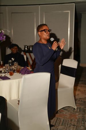 A speaker in a navy blue dress addresses an audience at a formal dinner event with white chairs and tables.