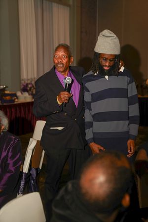 Two speakers share the microphone at a formal event with one wearing a suit and the other in casual attire.